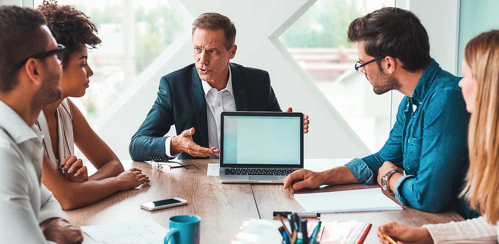 Analyzing results. Serious mature man in formal wear showing something on the digital tablet and discussing it with his multicultural team while sitting at the office table. Brainstorming